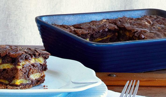 Fotografia de um pedaço da torta de banana em camadas em cima de um prato redondo e azul com um garfo ao lado, ao lado refratário azul com o restante da torta, tudo sobre bancada de madeira e fundo cinza.