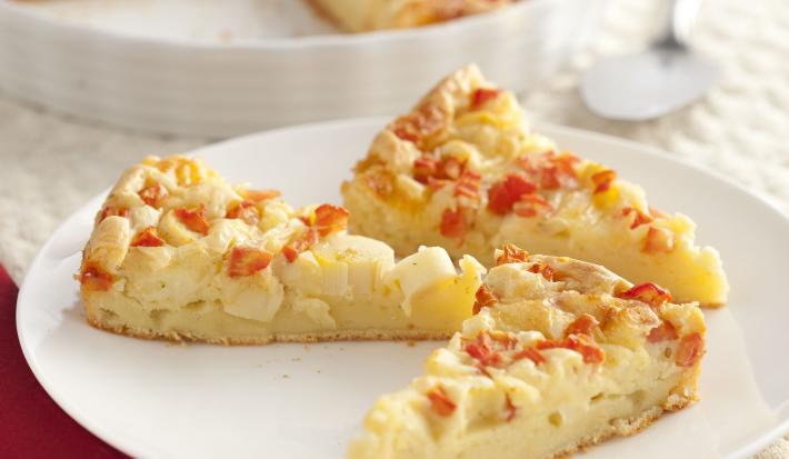Fotografia em tons de vermelho em uma bancada de madeira com um jogo americano bege, um pano vermelho, um prato branco raso com fatias da torta com legumes em cima dele. Ao fundo, um prato branco maior com a torta inteira.