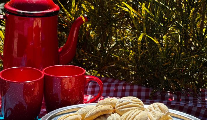 Fotografia de um recipiente fundo de alumínio com biscoitos amanteigados de leite moça. O recipiente está sobre um pano de mesa quadriculado de cor vermelho e branco. O recipiente está ao lado de duas xícaras e uma chaleira vermelha.