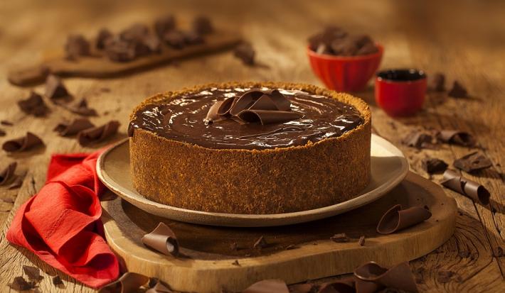 Fotografia em tons de marrom em uma bancada de madeira de cor marrom. Ao centro, uma tábua de madeira contendo o cheesecake e ao redor há pedaços de chocolates espalhados.