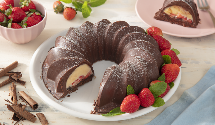Foto em tons claros da receita de bombom de morango servida em um prato de porcelana grande branco com uma porção de morangos. Ao lado, há raspas de chocolate meio amargo e ao fundo, um bowl rosa com morangos e um pedaço do bombom com um garfo prateado