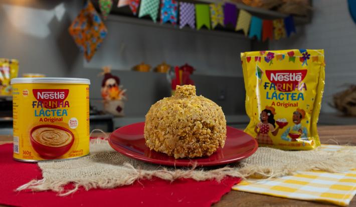 Fotografia de um prato pequeno, raso e vermelho apoiado em alguns panos de cor bege, um vermelho e outro amarelo quadriculado com branco. No prato tem uma receita de cuscuz com farinha láctea, ao lado do prato tem as embalagens da marca.