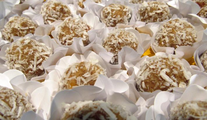 Fotografia de docinhos de beijinho com batata doce, passados no coco ralado e em forminhas de brigadeiro na cor branca, sobre uma mesa amarela.