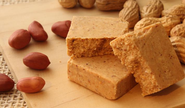 Fotografia em tons de caramelo com uma paçoca de formato retangular ao centro. Em volta da paçoca existe alguns amendoins