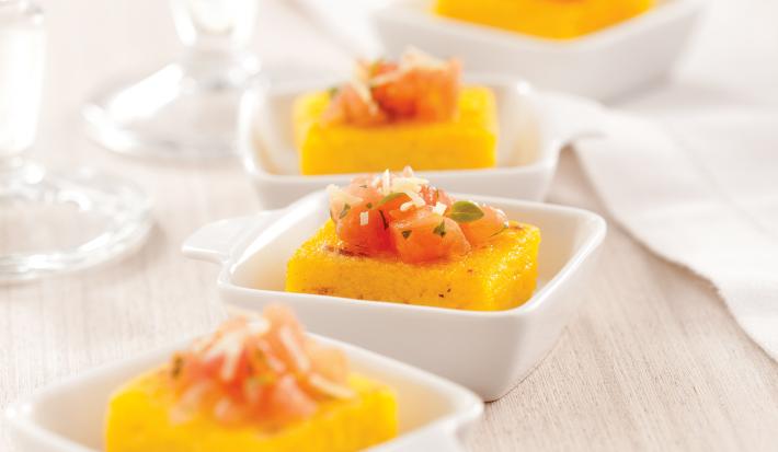 Fotografia em tons de laranja em uma bancada de madeira clara, um pano bege, quatro potinhos brancos pequenos com a polenta com tomate dentro de cada um.