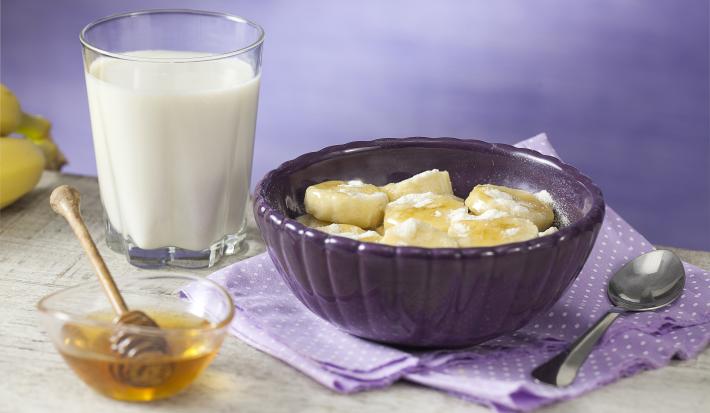Fotografia em tons de roxo em uma bancada de madeira com um pano roxo com bolinhas brancas, um pote grande e fundo roxo com as bananas cobertas com aveia e meia. Ao lado, um copo de leite, um potinho com o mel.