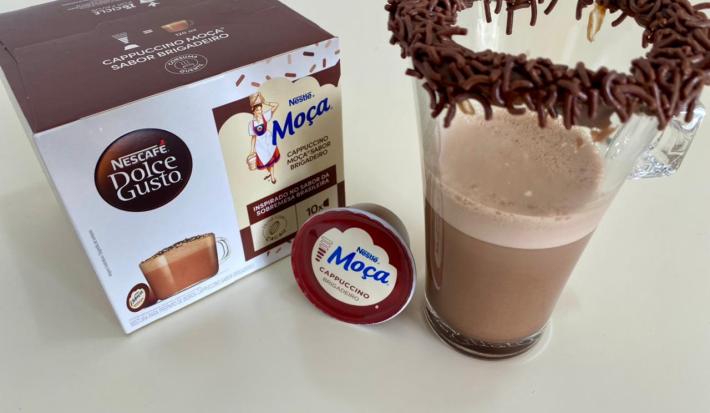 Fotografia de uma xícara de vidro com uma bebida de dolce gusto brigadeiro, com brigadeiro e granulado na borda. Ao lado tem uma cápsula de dolce gusto brigadeiro e a caixa com as cápsulas.