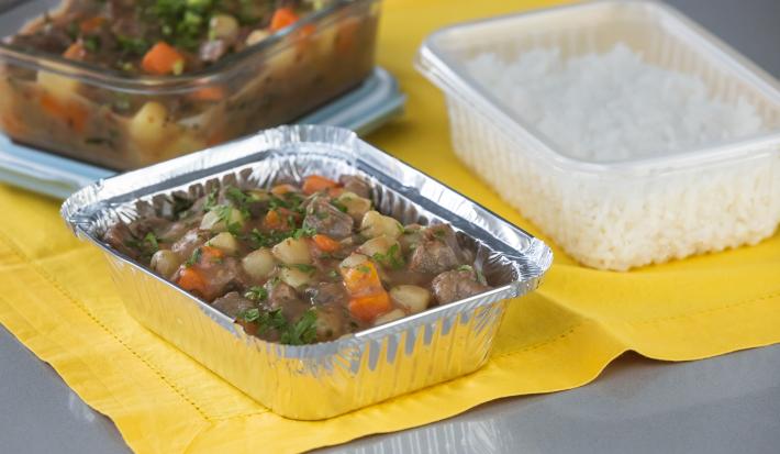 Foto da receita de picadinho de carne, servido em um recipiente de alumínio, sobre uma toalha amarela e acompanhado de arroz num pote de plástico