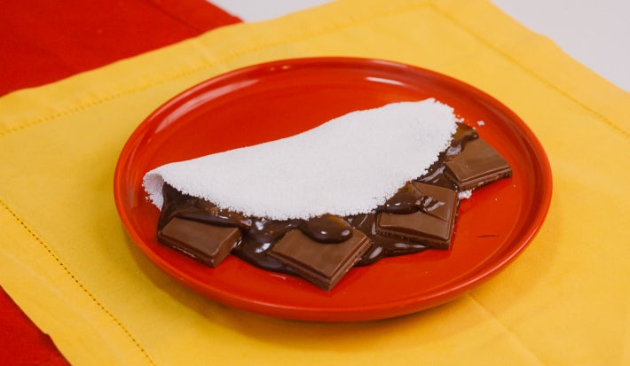 Tapioca dobrada recheada com creme de chocolate e pedaços de Baton, servida em prato vermelho.