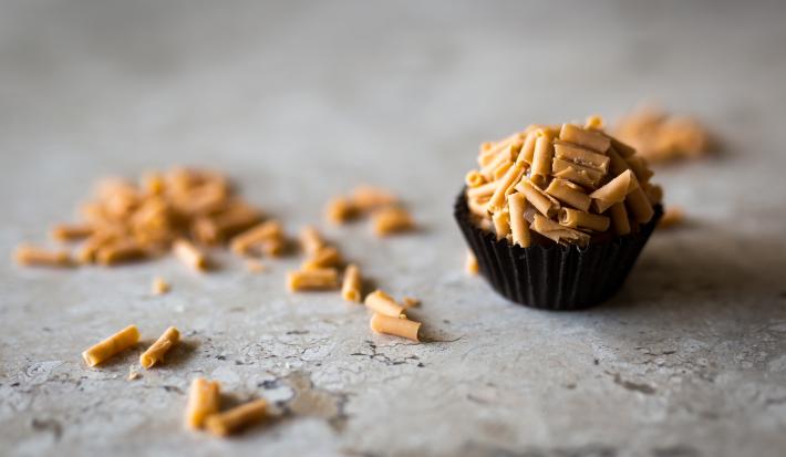 fotografia em tons de cinza, marrom e preto de uma bancada cinza vista de frente. Contém uma forminha preta com um brigadeiro dentro, por cima raspas de caramelo e na bancada contém as raspas espalhadas.