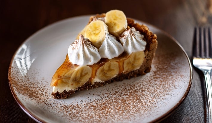 Em cima de uma mesa, fatia de banoffee com massa de biscoitos crocante, doce de leite, bananas em rodelas, decorado com chantilly cremoso, banana em rodelas e cacau em pó.