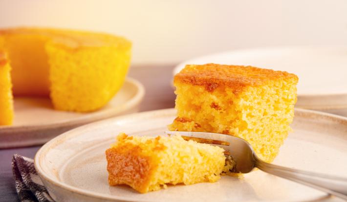 Fotografia de um prato grande de vidro com uma fatia grande de bolo de milho e um garfo cortando um pedaço. No fundo tem o bolo inteiro, sem essa fatia, sobre um apoio raso de cor clara.