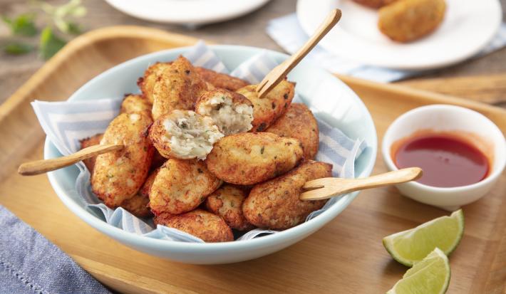 Fotografia em tons de bege em uma bancada de madeira clara, uma tábua de madeira, um pano azul, um prato redondo fundo com vários bolinhos de peixe, um potinho branco pequeno com molho de pimenta e pedaços de limão. Ao fundo, pratinhos brancos com bolinho