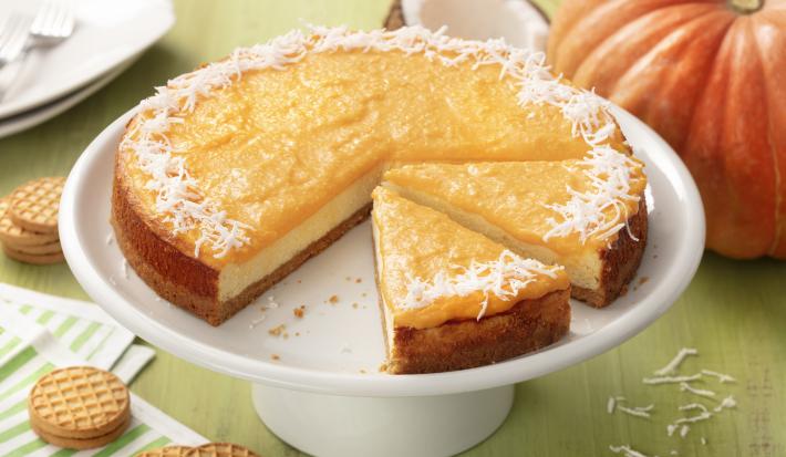 Fotografia em tons de verde e laranja de uma bancada de madeira verde com um paninho listrado verde e branco com biscoitos  e uma bailarina branca com o cheesecake de abóbora. Ao fundo uma abóbora, um coco e um prato quadrado branco.