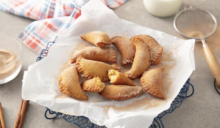 Foto da Receita de Pastel de Grão-de-bico. Observa-se um prato com papel absorvente e os pasteizinhos em cima polvilhados de canela e açúcar. Ao lado esquerdo, dois paus de canela e, ao direito, uma peneira com.