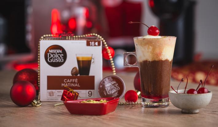Fotografia em tons de vermelho em uma bancada de madeira, uma xícara alta com o affogato natalino decorado com uma cereja. Ao fundo, enfeites e decoração de natal.