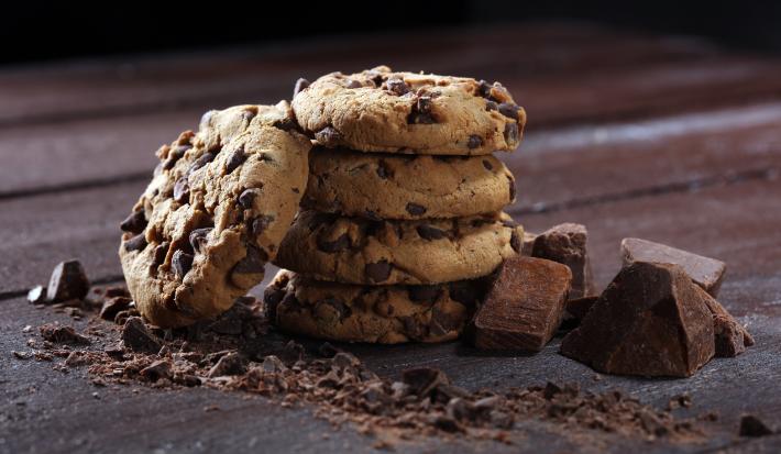 Fotografia de quatro cookies com gotas de chocolate, um sobre o outro. Encostado neles tem mais um cookie, ao lado de pedaços de chocolate, sobre uma mesa de madeira escura.