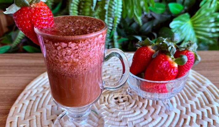 Fotografia em tons de rosa e marrom com uma caneca grande ao centro. Dentro da caneca existe uma bebida gelada a base de cápsulas de Dolce Gusto, e morangos congelados. Ao lado, existe um potinho com vários morangos.