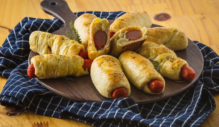 Foto aproximada de uma bancada sobre a qual está um pano azul quadriculado e uma tábua com a receita de enroladinho de salsicha nela.