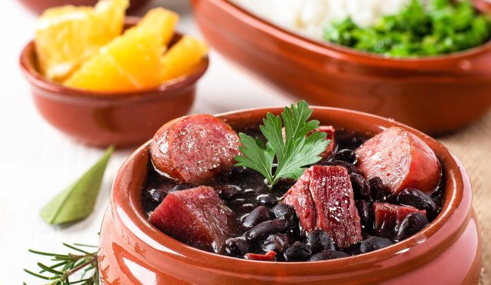 Fotografia de um recipiente médio marrom clássico com uma feijoada com caldo de bacon, linguiça e bacon com uma folha de salsinha. Ao fundo tem outros recipientes iguais, porém menores, com laranja, arroz, couve e farofa.