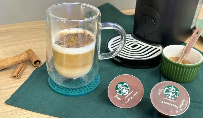 Foto da receita de Cappuccino Canela. Observa-se um copo transparente com o cappucino dentro, polvilhado de canela e chocolate em pó. Ao lado direito, cápsulas de Starbucks.