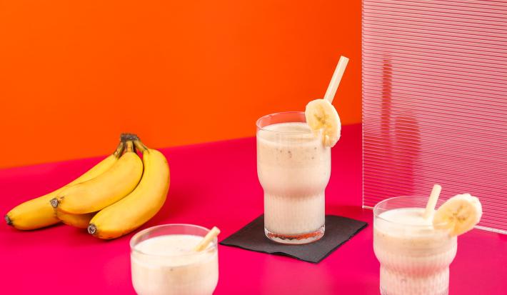 Fotografia de três copos de vidro com vitamina de banana e goiaba, e em cada copo, tem um canudo e uma rodela de banana decorando-o. Ao lado esquerdo, um cacho com quatro bananas, sobre uma mesa rosa com fundo laranja.
