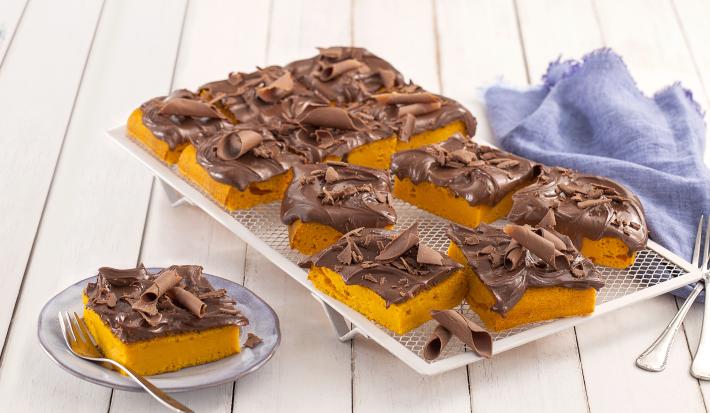Fotografia em tons de azul em uma bancada de madeira branca, um pano azul, um apoiador de grelha branco com vários pedaços do bolo de cenoura com cobertura de chocolate. Ao lado, um prato pequeno com uma fatia do bolo.