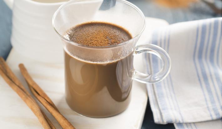 fotografia em tons de branco, marrom e azul tirada de uma xicara transparente com o Cappuccino dentro, e ao lado direito contém uma xicara branca e à frente canela.