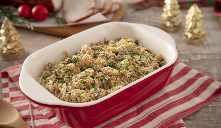 Foto da receita de Farofa de Bacon. Observa-se um refratário vermelho oval  com a farofa dentro, sobre um pano de prato listrado vermelho e branco. Itens de decoração natalinos enfeitam a foto.