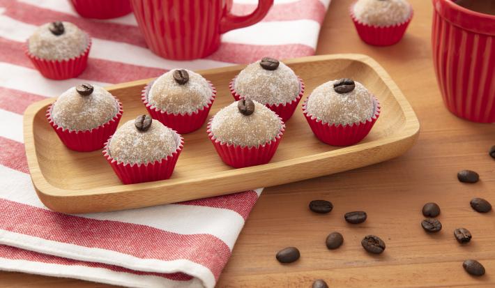 Fotografia em tons de vermelho de uma bancada de madeira, ao centro um pano listrado vermelho e branco e uma bandeja de madeira com seis brigadeiros em forminhas vermelhas, em cima de cada um deles um grão de café.