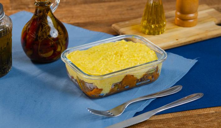 Fotografia de uma marmita pequena retangular com escondidinho de mandioquinha com picadinho de carne abobrinha. A marmita está sobre dois panos azuis sobrepostos, ao lado de talheres e uma tábua de madeira de tom claro.