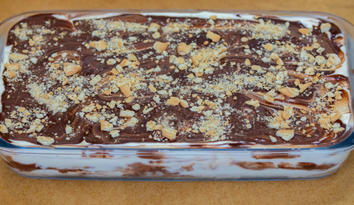 Fotografia mostra uma travessa retangular trasnparente servindo um creme branco e de chocolate. Pedaços de biscoito triturado por cima