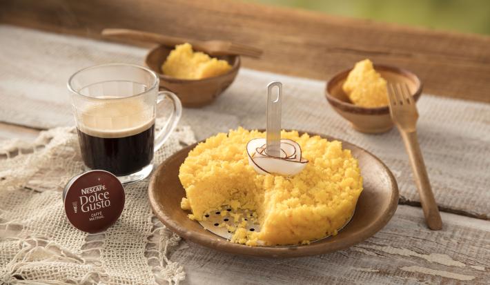 Fotografia em tons de amarelo, branco e marrom de uma bancada com um pano rendado, uma cápsula de Dolce Gusto e uma xícara transparente com café. Ao lado um prato pequeno redondo com o cuscuz e tiras de coco. Ao fundo, dois potinhos com cuscuz e 2 garfos. Fotografia em tons de amarelo, branco e marrom de uma bancada com um pano rendado, uma cápsula de Dolce Gusto e uma xícara transparente com café. Ao lado um prato pequeno redondo com o cuscuz e tiras de coco. Ao fundo, dois potinhos com cuscuz e 2 garfos.