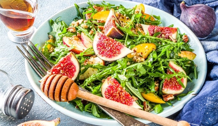 Em cima de uma mesa, prato com salada com mix de folhas, frutas em cubos e figo.