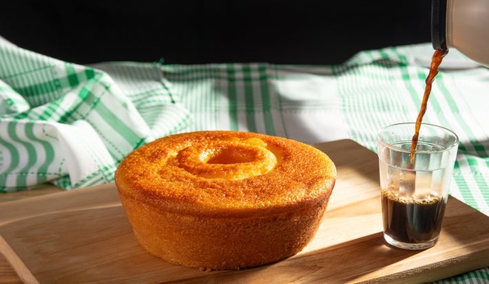 Um bolo de cenoura inteiro, com formato redondo e com furo no meio, está sobre uma tábua de madeira. Ao lado, um copo está sendo servido com café. O fundo tem um pano de prato verde e branco.