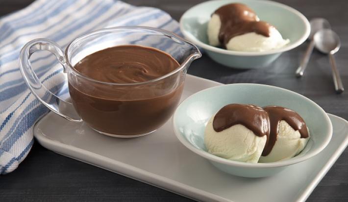 Foto em tons de marrom da receita de creme de chocolate diferente servida em uma xícara de vidro e ao lado há um sorvete servido com o creme por cima, isso tudo sobre uma base de porcelana branca. Ao fundo mais uma porção do sorvete com duas colheres.