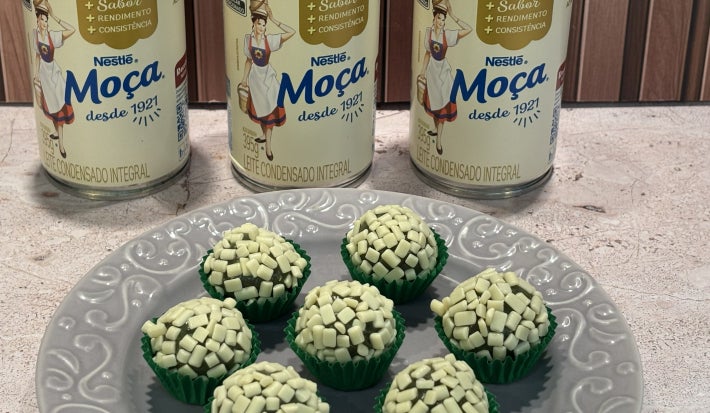 Docinhos verdes com granulado branco, em forminhas verdes sobre prato cinza, com latas de Leite Moça ao fundo.