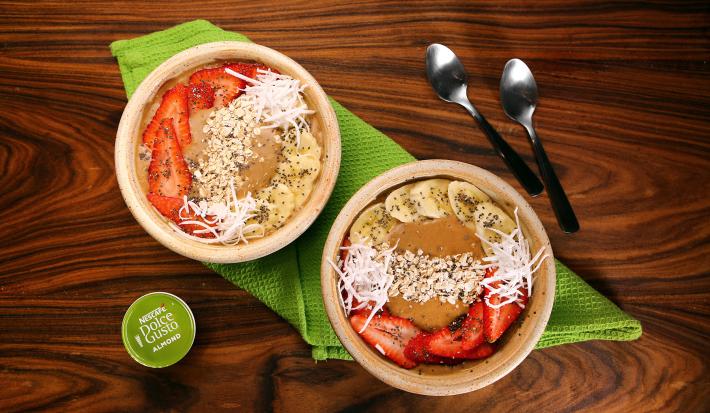 Fotografia em tons de verde em uma bancada de madeira escura, um pano verde, dois bowls bege com morango, banana e lascas de coco e duas colheres ao lado.