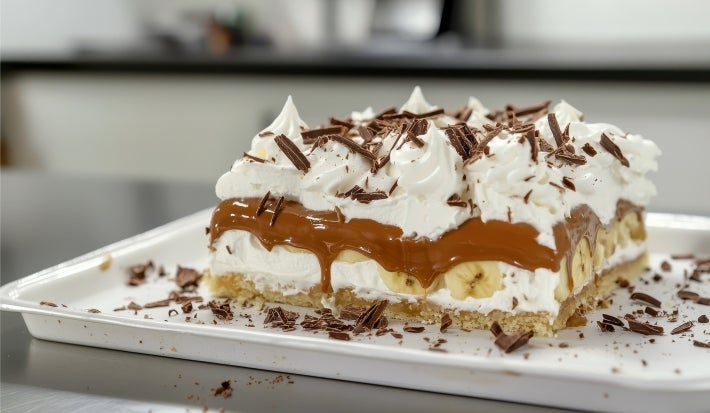 Em cima de uma mesa, prato com fatia de banoffee com massa crocante, chantilly com doce de leite e bananas em rodelas, decorado com chantilly e raspas de chocolate.