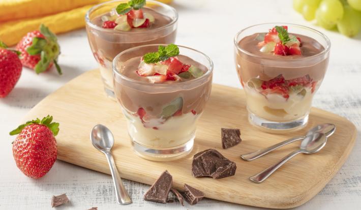 Fotografia em tons de amarelo em uma bancada cinza com uma tábua de madeira no centro e três potinhos de vidro com o bombom de chocolate, creme e frutas. Ao lado, pedaços de chocolate, morangos e um cacho de uvas espalhados.