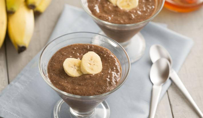 Fotografia em tons de cinza, marrom, amarelo e azul de uma bancada cinza vista de cima, ao centro um pano azul com duas taças transparente ambas contém mousse de chocolate e duas fatias de banana. Ao fundo um cacho de banana, mel e ao lado duas colheres