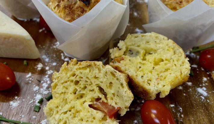 Foto da Receita de Muffin de Pizza. Observa-se um muffin cortado ao meio em cima de uma tábua de madeira. Tomates cereja e queijo decoram a foto também