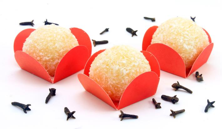 Fotografia de três unidades de beijinho com gelatina dentro de embalagens vermelhas sobre uma mesa branca, com cravos espalhados.