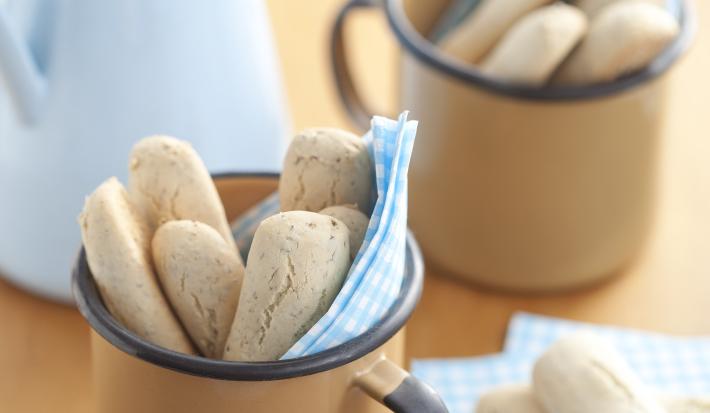 Fotografia em tons de azul em uma mesa de madeira com um paninho azul claro com bolinhas brancas, ao centro uma xícara bege com os biscoitinhos nutritivos. Ao fundo, uma jarra azul claro.