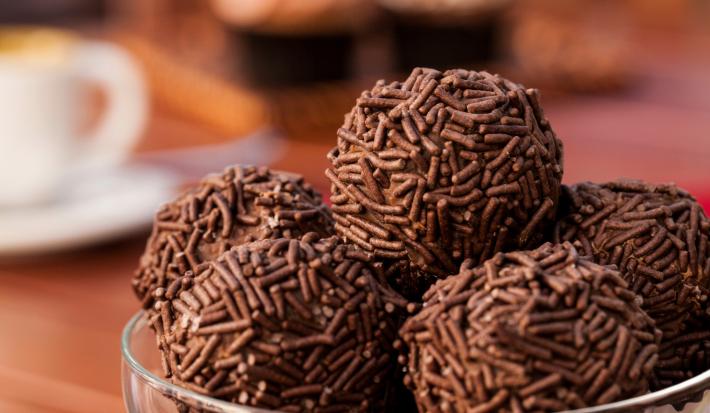Fotografia de vários brigadeiros enrolados no granulado e recheados com panettone. Os brigadeiros estão em um recipiente fundo de vidro, mas que só aparece na borda, e está sobre uma mesa de madeira.