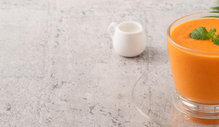 Fotografia em tons claros de uma vitamina laranja com folhas de hortelã por cima em um copo de vidro, apoiado sobre um prato pequeno de vidro. Ao fundo, um pote pequeno branco de vidro, sobre uma mesa em tons de cinza.