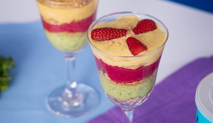 Fotografia em tons claros de duas taças de vidro com os cremes coloridos intercalados com o biscoito nesfit. As taças estão sobre dois panis, azul e roxo, ao lado tem a lata de ascenda e três morangos inteiros.
