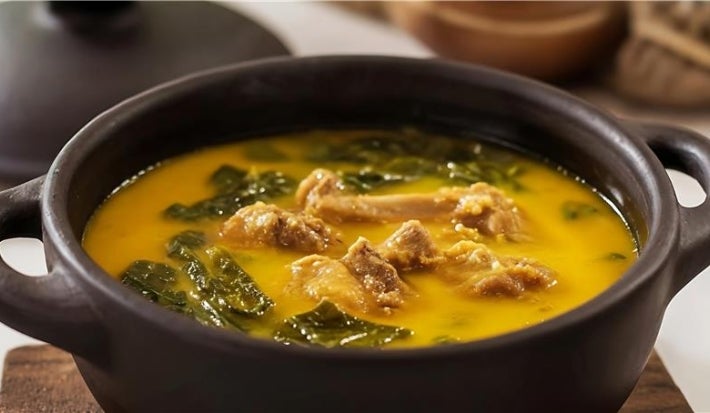 Receita de frango servido no caldo de tucupi com folhas de jambu. Uma foto bem aproximada que mostra uma panela de barro preta com tudo dentro.