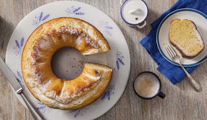 Fotografia em tons de cinza, branco e azul de uma bancada cinza e um prato banco com detalhes azuis, sobre ele o bolo de milho. Ao lado duas xícaras de café com leite e um paninho azul com um prato branco de bordas azuis com uma fatia do bolo e um garfo.