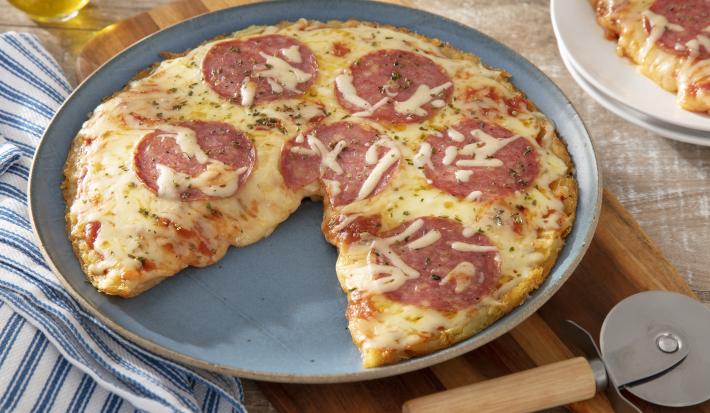Foto da receita de pizza caseira com massa de batata com rodelas de salame por cima sobre uma base circular de cerâmica com um cortador de pizza prateado ao lado sobre uma tábua de madeira. Ao lado há um paninho azul listrado e um vidro de azeite